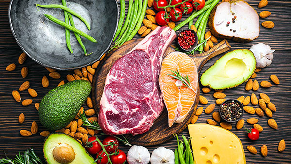 assortment of keto food on a tabletjop including steak, chicken, avocado, and various nuts